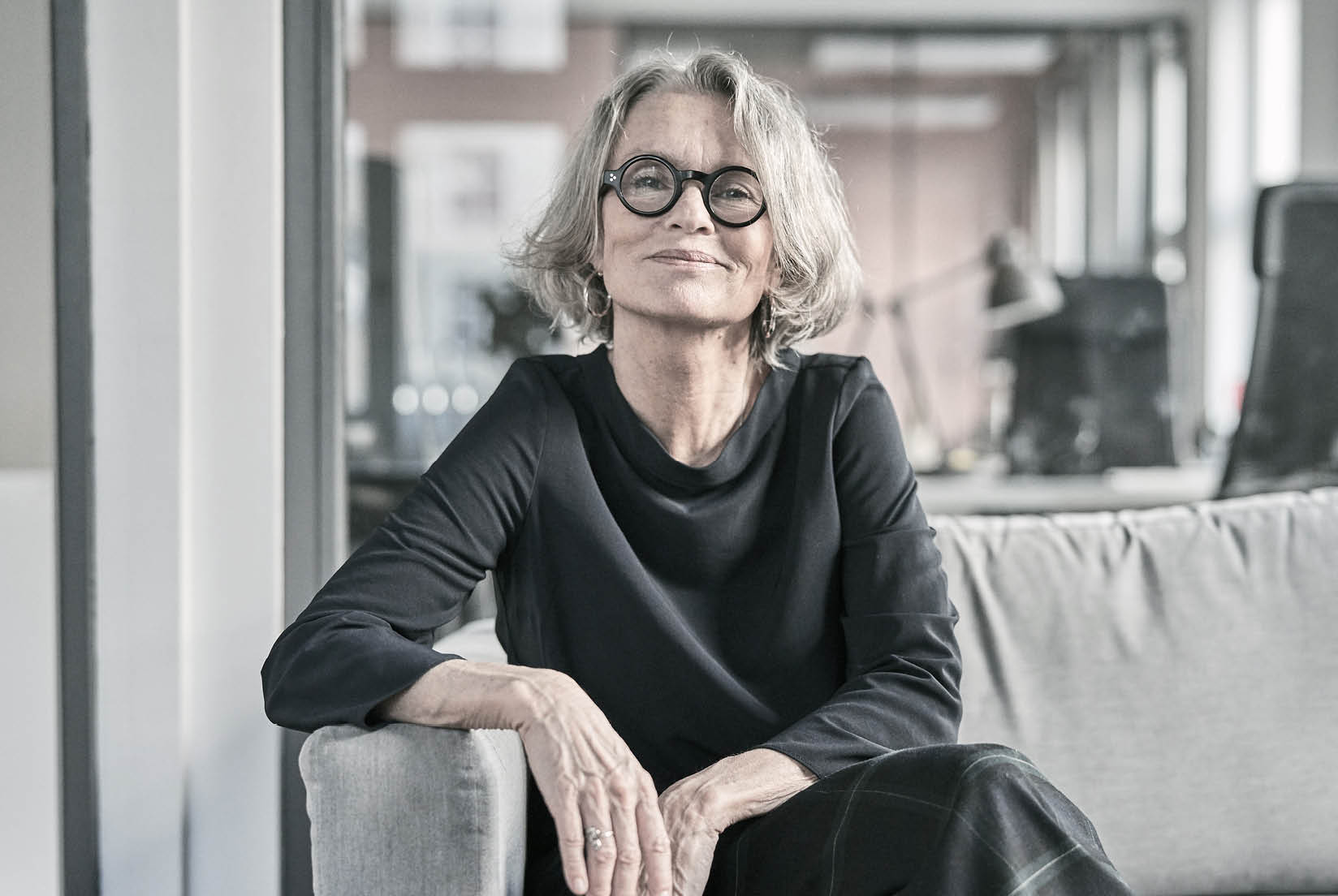 A mature woman seated on a sofa with legs crossed, one elbow resting on the sofa arm and the other hand on her tights. She’s centered in the frame, looking directly at the camera with a gentle smile. The image is in grey tones with low contrast.