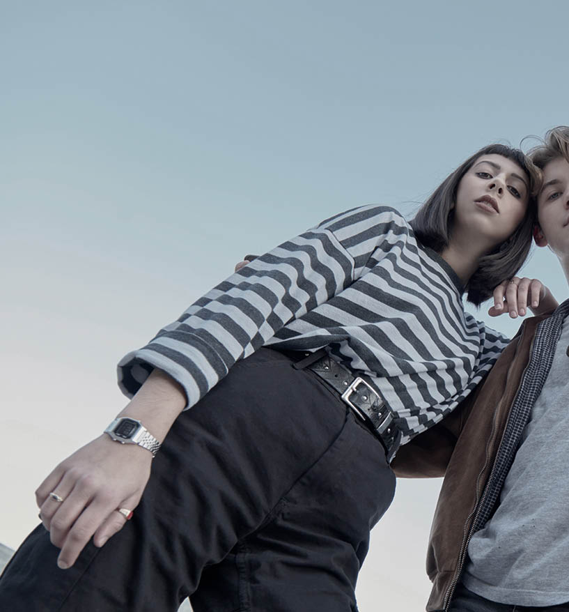 Log/raw footage showing a young girl resting her arm on a young boy’s shoulder, while he has his arm around her lower back. They are standing close together and looking down toward the camera, which is positioned below them.