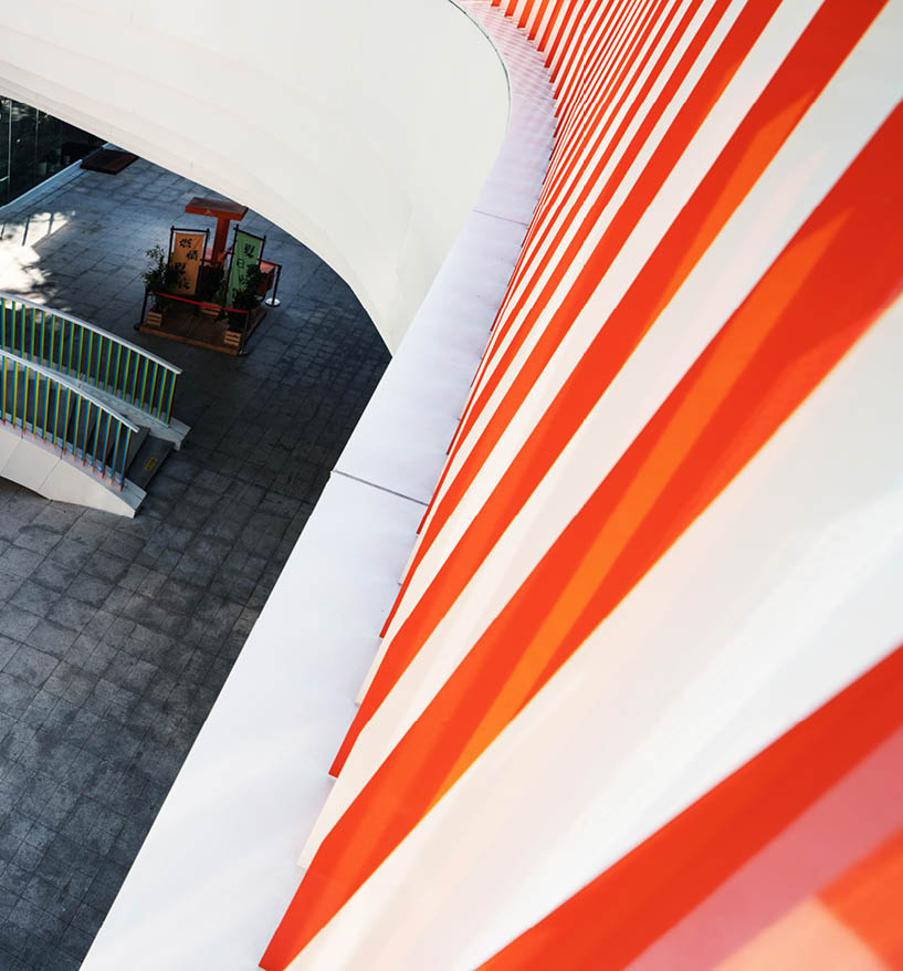 Photo taken from above of an outdoor white and orange striped wall, with sunlight streaming in. The shaded end of a stairwell is visible on the floor below.